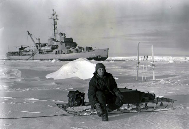 Man sits on ice with ship in background.