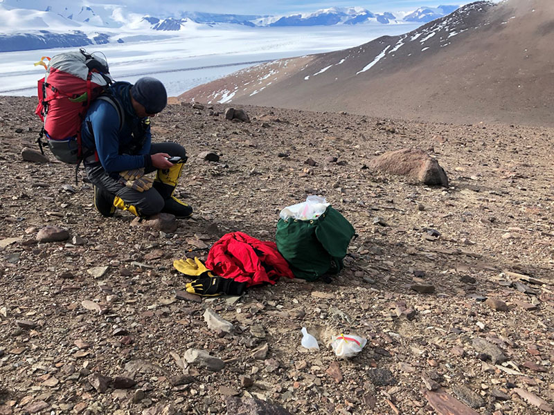 Mountaineer Geoff Schellens kneels and takes a GPS measurement where the team collected a soil sample, with the Transantarctic Mountains in the background