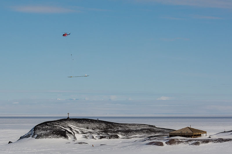With SkyTEM slung underneath, a Bell 212 helicopter flies past the Discovery historic hut on Hut Point Peninsula