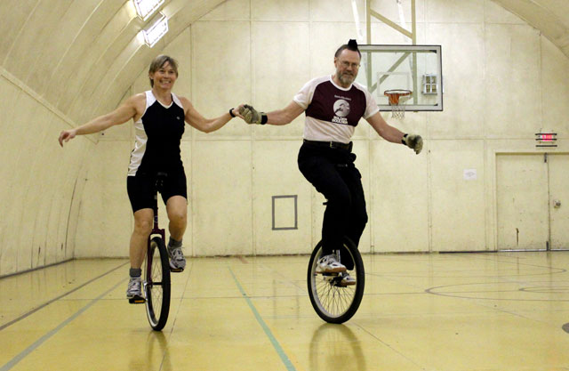 Two people on unicycles in a gym.