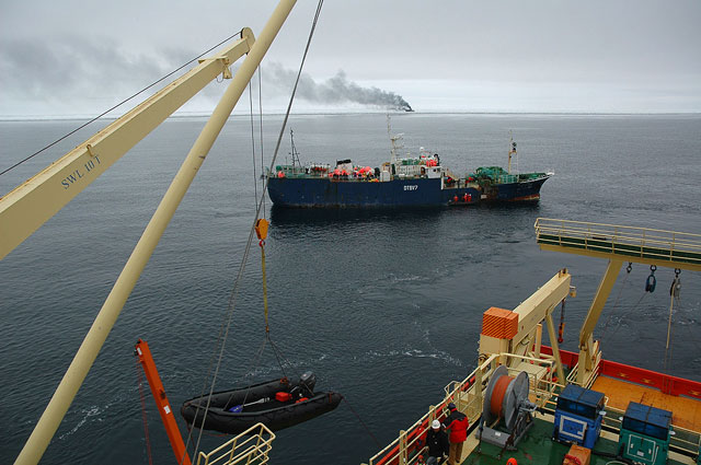 A ship crane unloads a Zodiac with two ships in background.