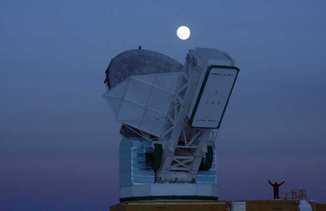 Person raises arms next to telescope.