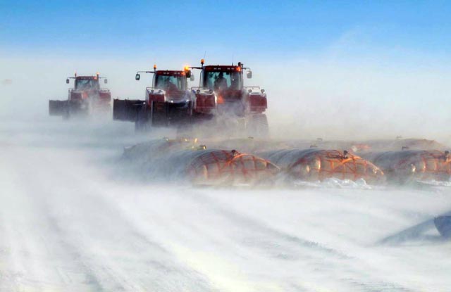 Snow blows around a line of tractors and bladders.