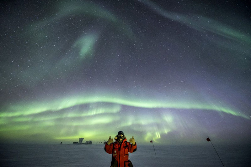 Robert Schwarz gives the thumbs up under bright auroras, with the South Pole telescope in the background