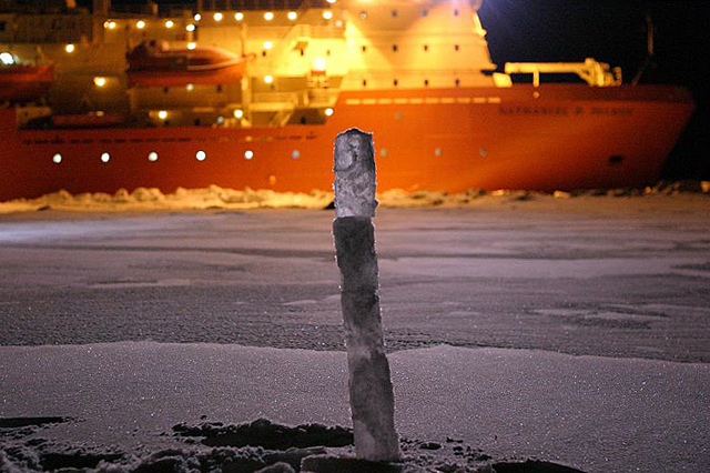 Cylinder of ice sticks up with ship in background.