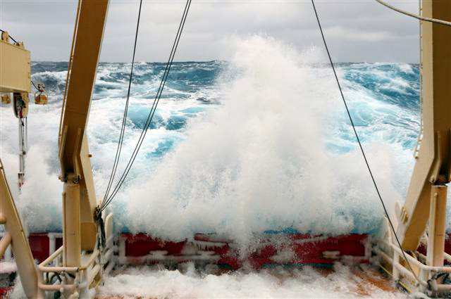 Waves crash over stern of ship.