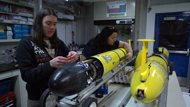 Two people assemble torpedo-shaped robots.