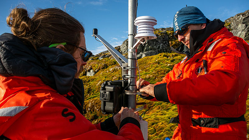 Ryan Newell (left) and David Beilman install sensors onto a small weather station near Palmer Station to collect data about the weather conditions that moss thrives in