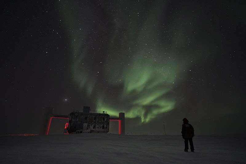 A lone figure stands out in the Antarctic Winter, watching the aurora shine over the IceCube laboratory