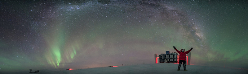 A station resident stands outside in front of the IceCube laboratory with the Milky Way and southern lights overhead